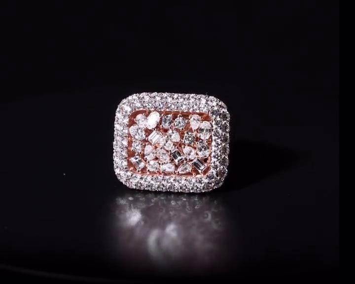 May include: A square-shaped ring with a rose gold band and a cluster of diamonds. The ring's face is covered in small, clear diamonds, with a border of larger diamonds. The center features a mix of diamond shapes, creating a sparkling effect. The ring is set against a dark background.