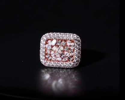 May include: A square-shaped ring with a rose gold band and a cluster of diamonds. The ring's face is covered in small, clear diamonds, with a border of larger diamonds. The center features a mix of diamond shapes, creating a sparkling effect. The ring is set against a dark background.