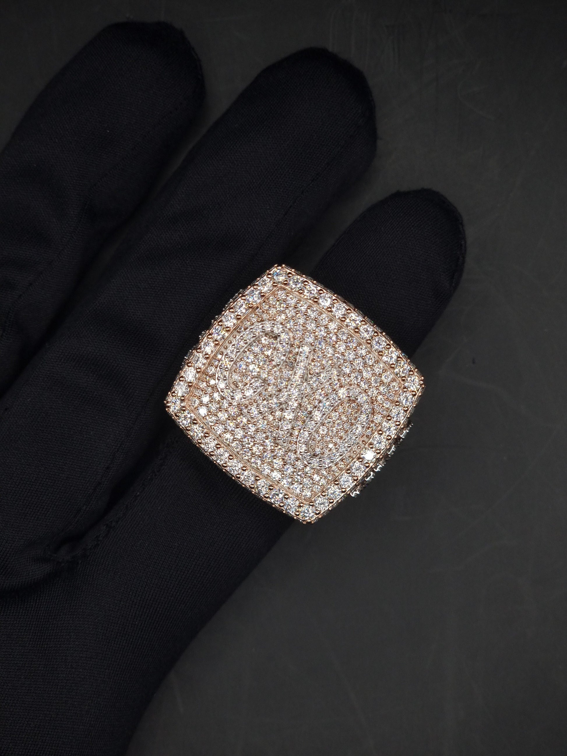 May include: A square shaped ring with a large, faceted, clear stone set in a rose gold setting. The stone is surrounded by smaller, clear stones.