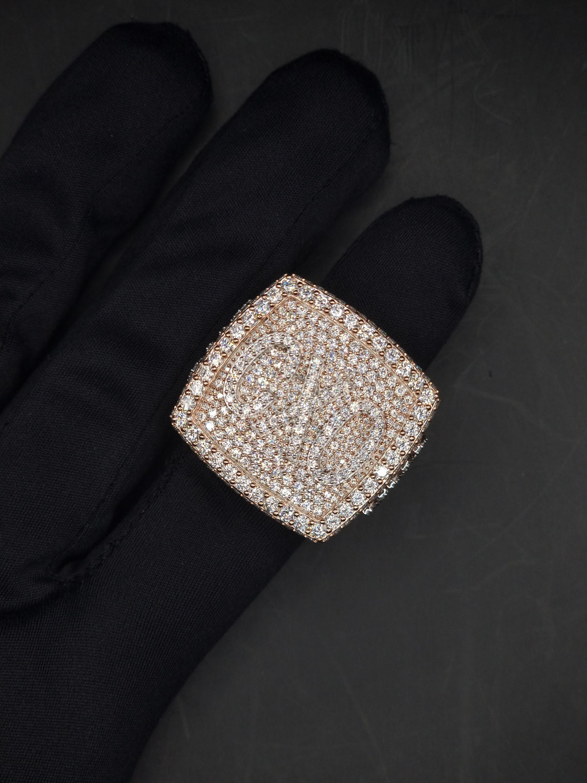 May include: A square shaped ring with a large, faceted, clear stone set in a rose gold setting. The stone is surrounded by smaller, clear stones.