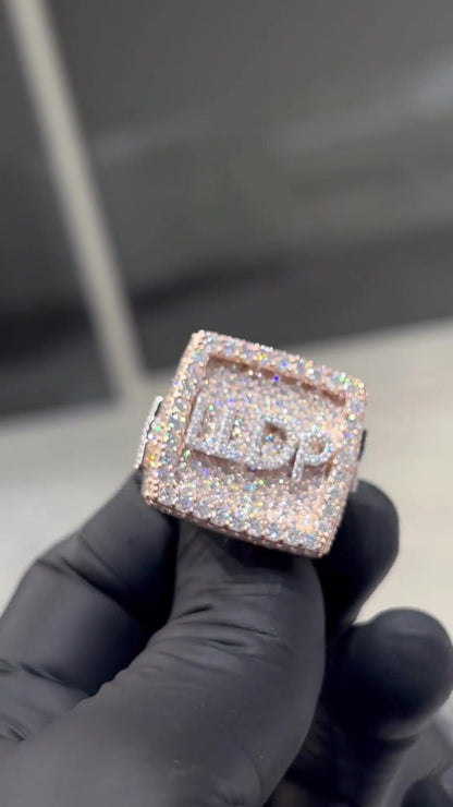 May include: A square-shaped ring with a rose gold band and a square-shaped center piece. The center piece is covered in small, clear diamonds and has the letters 'L.D.P.' in the center.