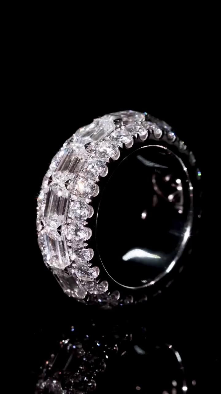 May include: A diamond ring with a black background. The ring is round with multiple rows of clear, sparkling diamonds. The diamonds are set closely together, creating a continuous band of brilliance. The ring's band is a dark, reflective material, enhancing the sparkle of the diamonds.