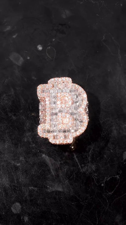 May include: A diamond-encrusted ring in the shape of the letter 'B'. The ring is made of rose gold and features a pave setting of small, clear diamonds. The diamonds are arranged to create the shape of the letter, with a slightly rounded top and a thicker base. The ring is displayed on a dark, textured surface.