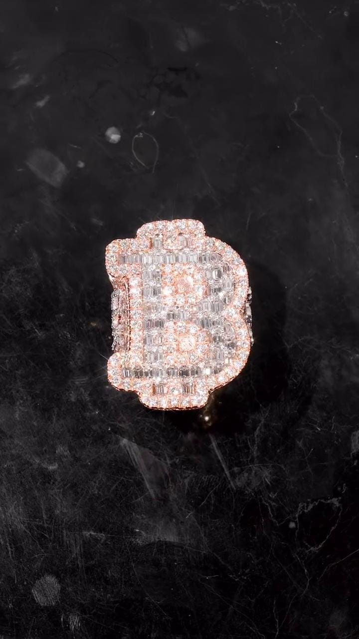 May include: A diamond-encrusted ring in the shape of the letter 'B'. The ring is made of rose gold and features a pave setting of small, clear diamonds. The diamonds are arranged to create the shape of the letter, with a slightly rounded top and a thicker base. The ring is displayed on a dark, textured surface.