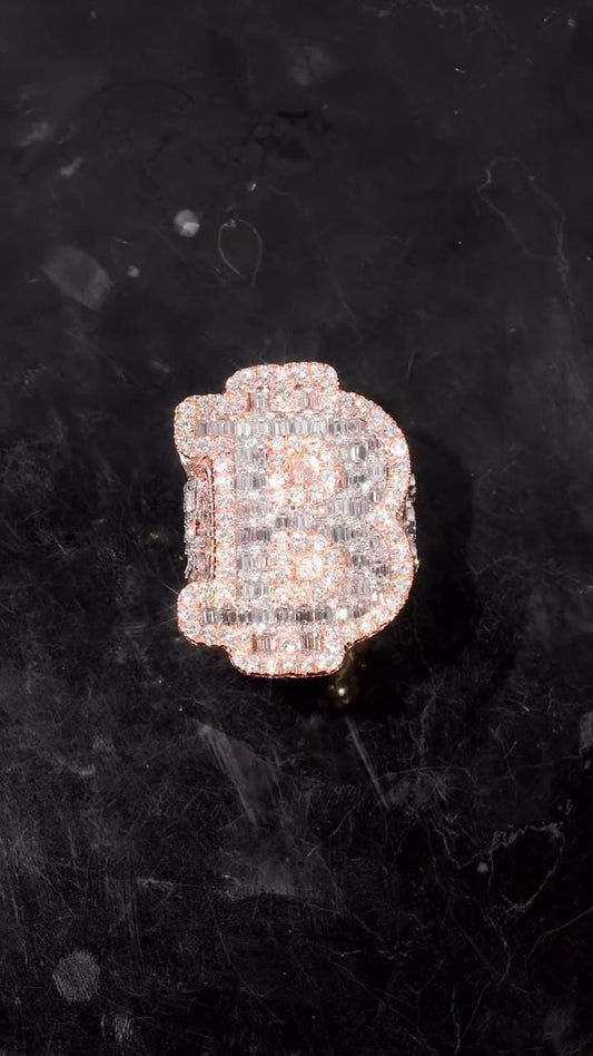 May include: A diamond-encrusted ring in the shape of the letter 'B'. The ring is made of rose gold and features a pave setting of small, clear diamonds. The diamonds are arranged to create the shape of the letter, with a slightly rounded top and a thicker base. The ring is displayed on a dark, textured surface.