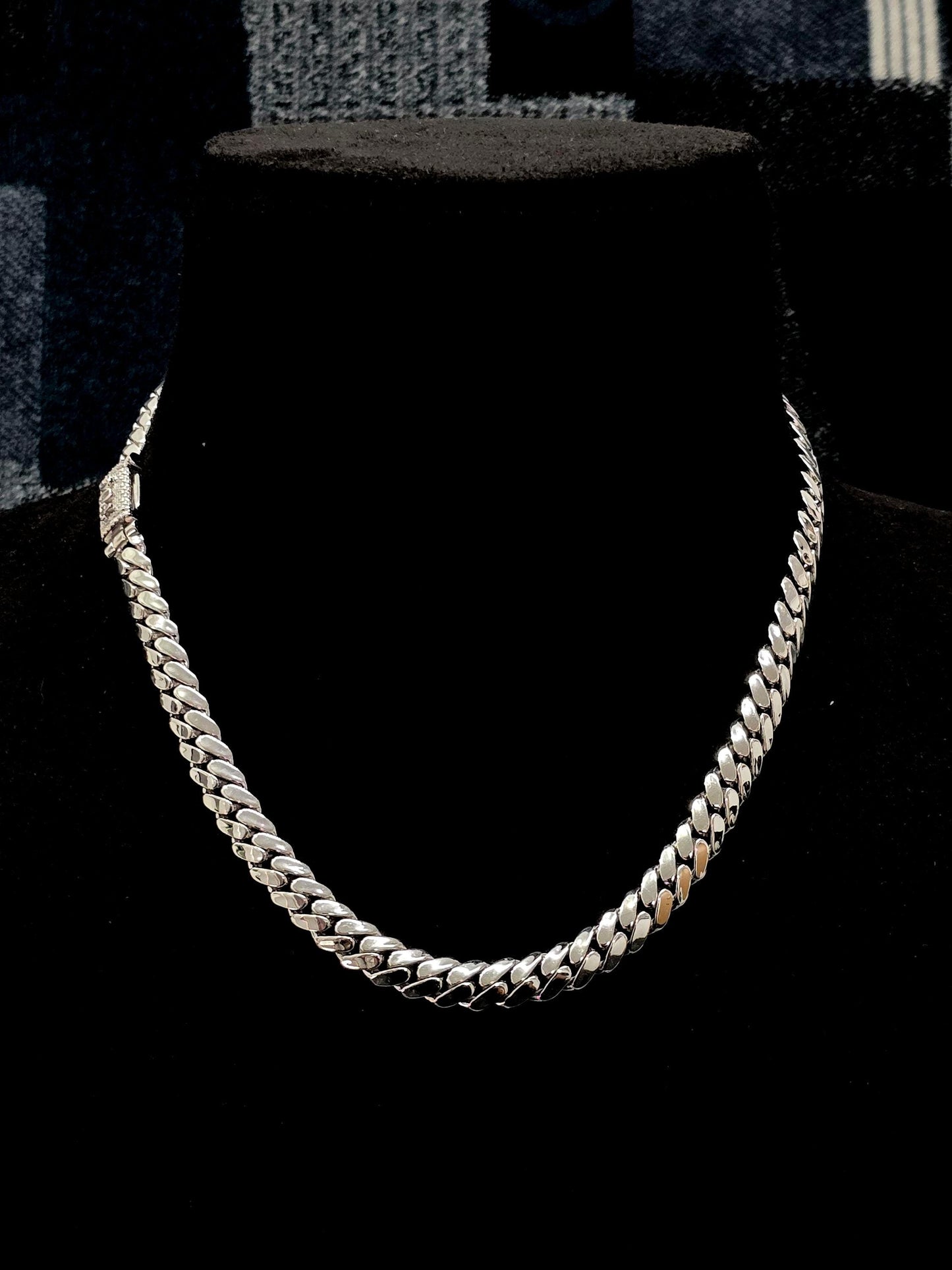 May include: A silver-colored curb chain necklace displayed on a black mannequin. The necklace has a thick, flat link design and a secure clasp. The background is a dark, solid color.