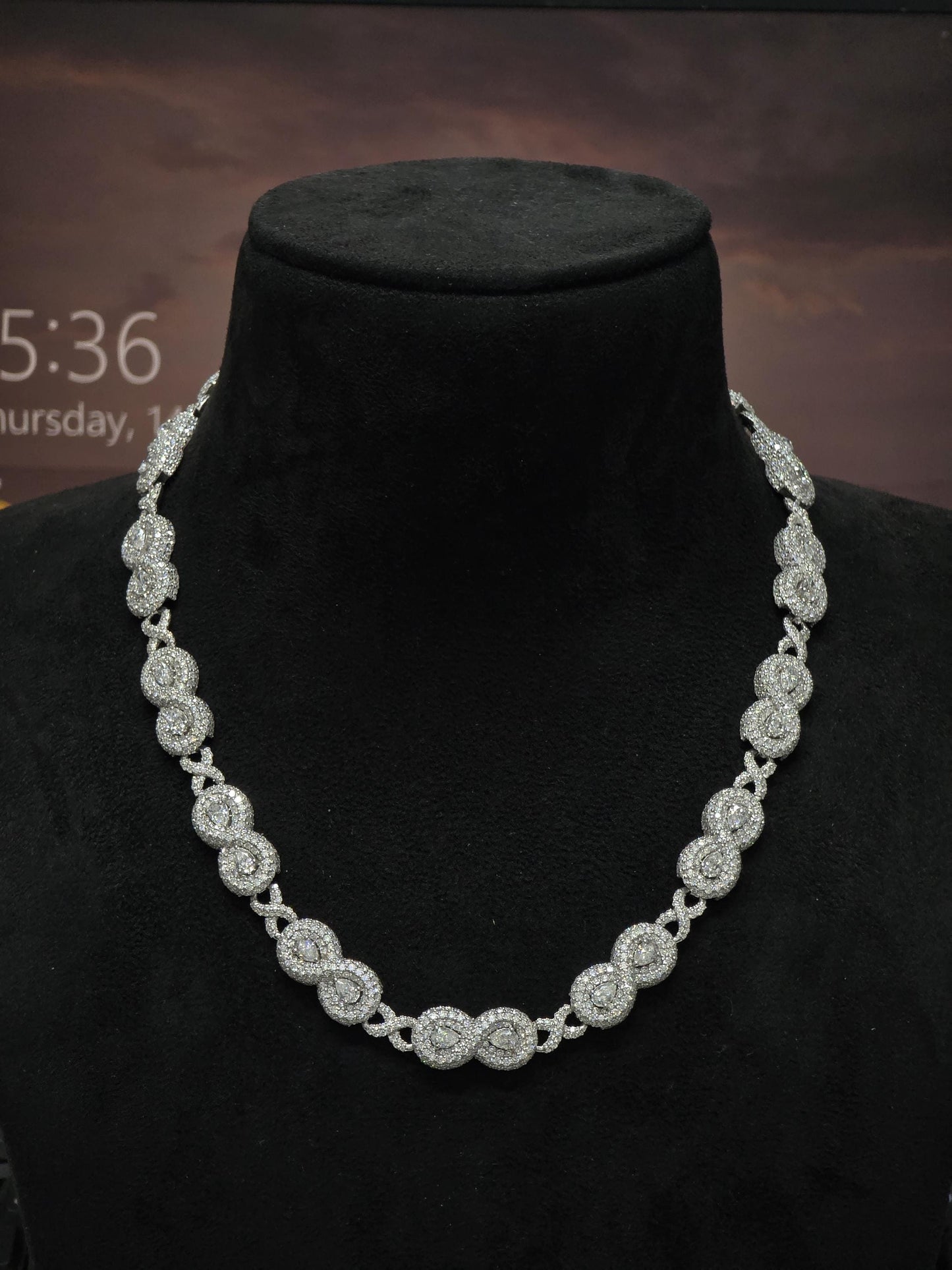 May include: A sparkling silver necklace with a repeating pattern of circular and X-shaped links. The necklace is displayed on a black mannequin, showcasing its intricate design and brilliant shine. The necklace is a statement piece.
